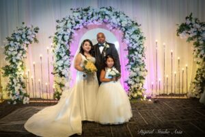 Bride, groom, and flower girl posing together under a floral wedding arch with soft candle lighting at Oasis Convention Centre.