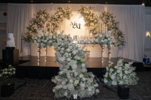 Elegant white wedding stage at Oasis Convention Centre decorated with cascading floral arrangements, candles, and a monogram backdrop.